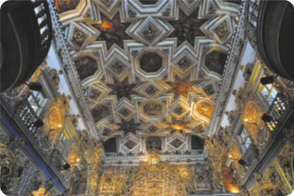 Fotografia do interior de uma igreja de arquitetura barroca, com as paredes revestidas com ornamentos dourados e o teto repleto de ornamentos com formas geométricas e pinturas.