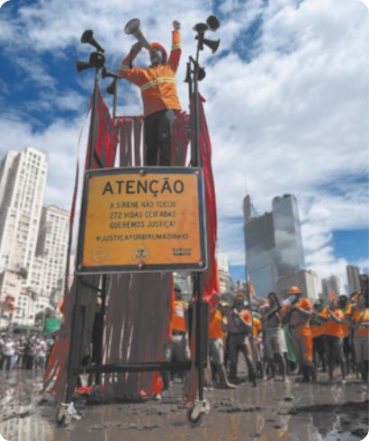Fotografia de um artista em uma performance em pé sobre um andaime em uma via pública. Ele está falando com um megafone na mão, usando um capacete e um uniforme, com o braço erguido para o alto. Uma placa no andaime diz: 'Atenção. A sirene não tocou. Duzentas e setenta e duas vidas ceifadas. Queremos justiça! # Justiça por Brumadinho'. O piso sob o andaime está coberto com água e lama, e há um grupo de manifestantes uniformizados ao lado do andaime.