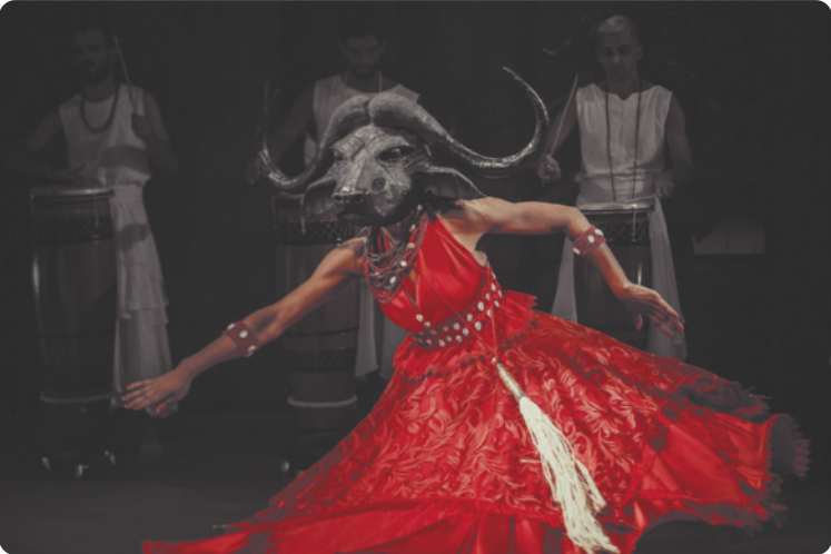 Fotografia de uma mulher dançando, de vestido, usando uma máscara com a forma da cabeça de um búfalo, com grandes chifres. Ao fundo há homens de branco tocando atabaques.