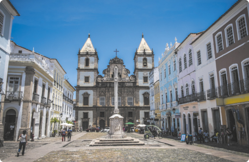 Fotografia de uma praça em frente a uma igreja e edifícios baixos de arquitetura barroca característica do período colonial brasileiro. No centro da praça há um monumento em forma de poste, erguido sobre três degraus de escadaria e um pedestal.