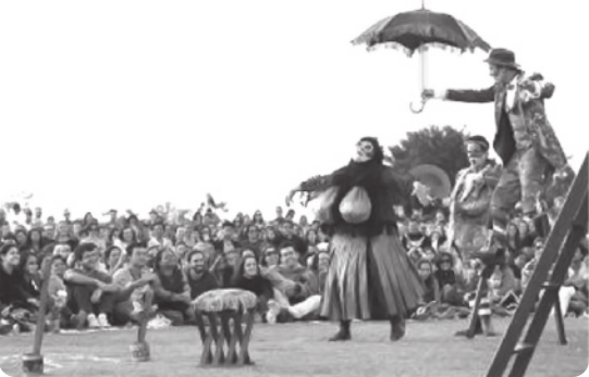 Fotografia em preto e branco de atores ao ar livre em um gramado, encenando uma peça. Um deles está de chapéu, andando sobre pernas de pau, segurando um guarda-chuva; outro está usando um leque; e há uma pessoa de saia, com uma máscara e bolsas penduradas em frente ao peito, caminhando com os braços abertos. Há uma plateia de pessoas sentadas no chão assistindo, e ao fundo uma fileira de pessoas assistindo em pé.