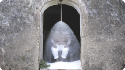 Fotografia de um homem sentado no interior de um engenho de açúcar, em frente à porta aberta, com uma cascata de açúcar caindo em sua cabeça, de modo que se forma uma espécie de véu branco sobre seu corpo.