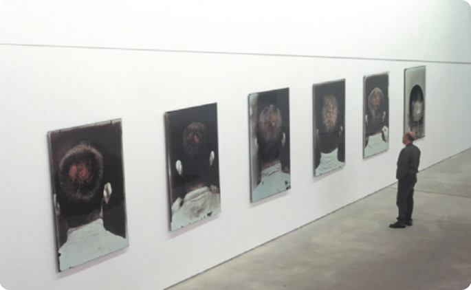 Fotografia de um espaço expositivo em que um homem observa um dos seis quadros que estão expostos na parede. Os quadros contêm fotografias com retratos em grandes formatos de pessoas de costas, com os cabelos raspados.