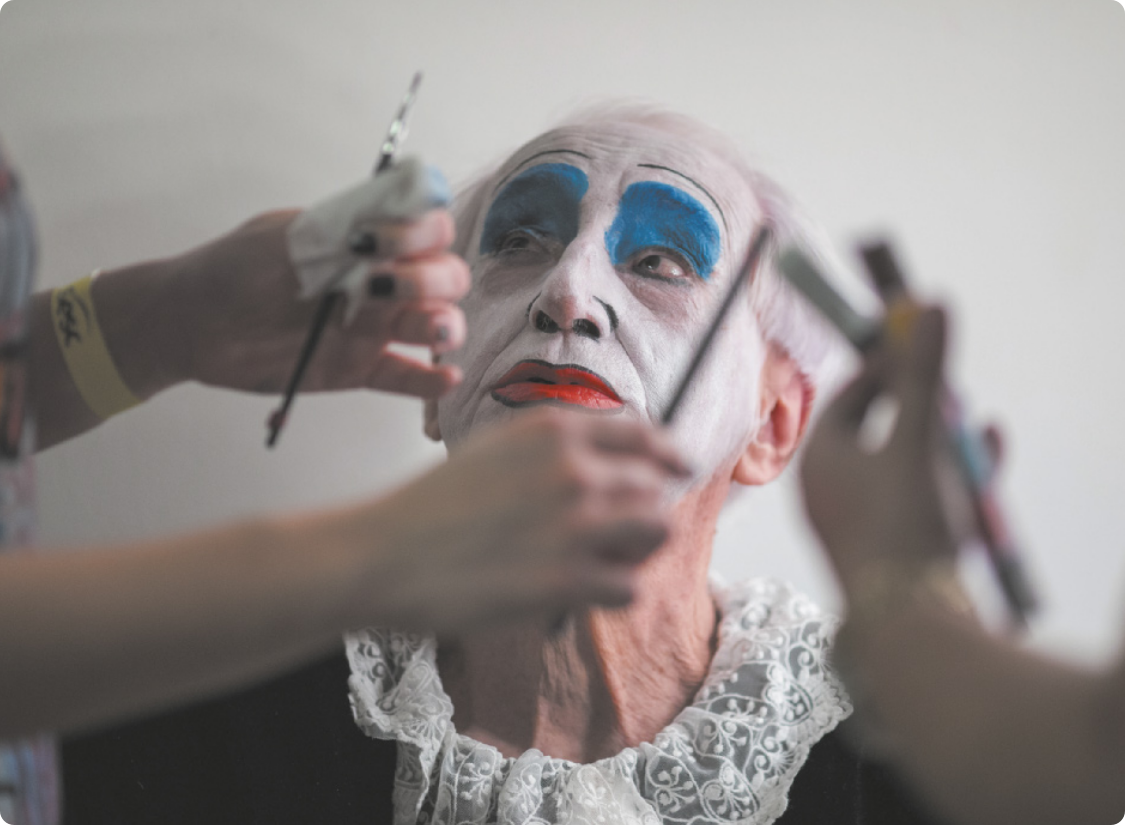 Fotografia de Zé Celso. Ele é um homem idoso, com cabelos grisalhos e o rosto pintado à maneira de um palhaço. Ele está vestido com uma gola de rendas, e olha para o alto enquanto pessoas pintam o rosto dele.