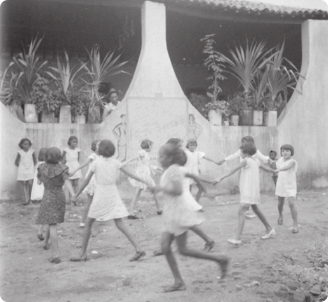 Fotografia em preto e branco de um grupo de meninas dançando em roda, de mãos dadas, em frente à varanda de uma casa.
