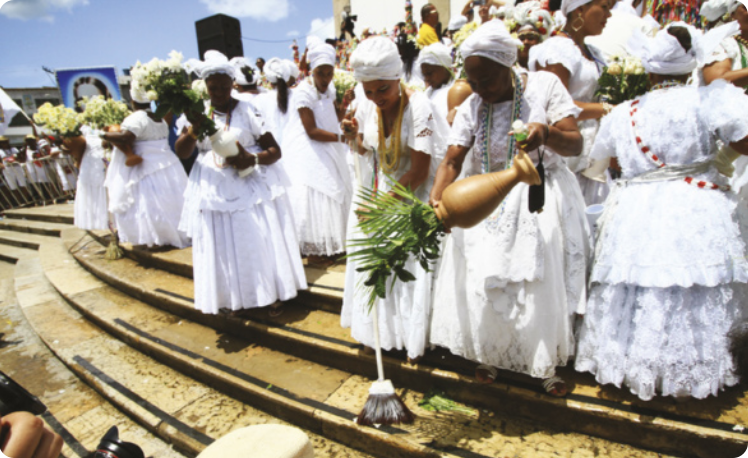 Fotografia de um grupo de mulheres com vestidos brancos e lenços na cabeça em uma escadaria, segurando vasos com buquês de flores. Uma delas está lavando o chão com uma vassoura.