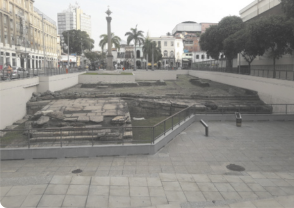 Fotografia de uma praça com parte de um calçamento antigo preservada, cercada por barras de ferro. Ao fundo há um poste, árvores e edifícios de alturas diversas.