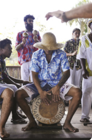 Fotografia de um músico tocando um tambor grande, sentado sobre ele, com outros músicos ao fundo, tocando chocalhos e um clarinete.