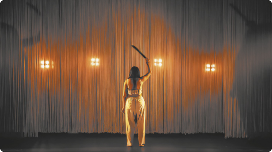 Fotografia de Zahy Tentehar. Ela está em pé, de costas, em um palco, erguendo um facão para o alto com uma das mãos.