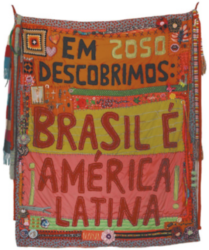 Fotografia de um tecido com bordados de tecidos e padrões diversos, além de formas abstratas e geométricas coloridas. No tecido está bordada a frase: 'Em 2050 descobrimos: Brasil é América Latina'.