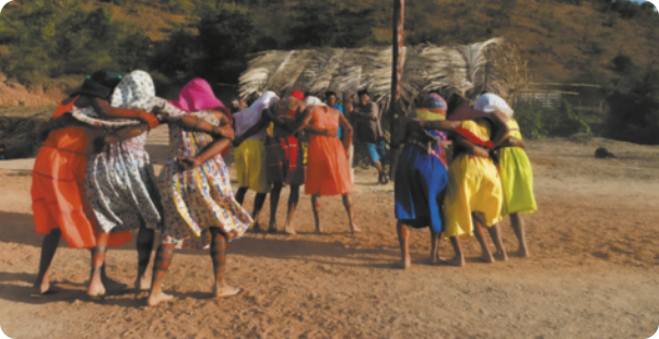Imagem de filme em que mulheres indígenas com lenços na cabeça e vestidos coloridos dançam abraçadas, em trios, sobre o solo de areia de uma aldeia.