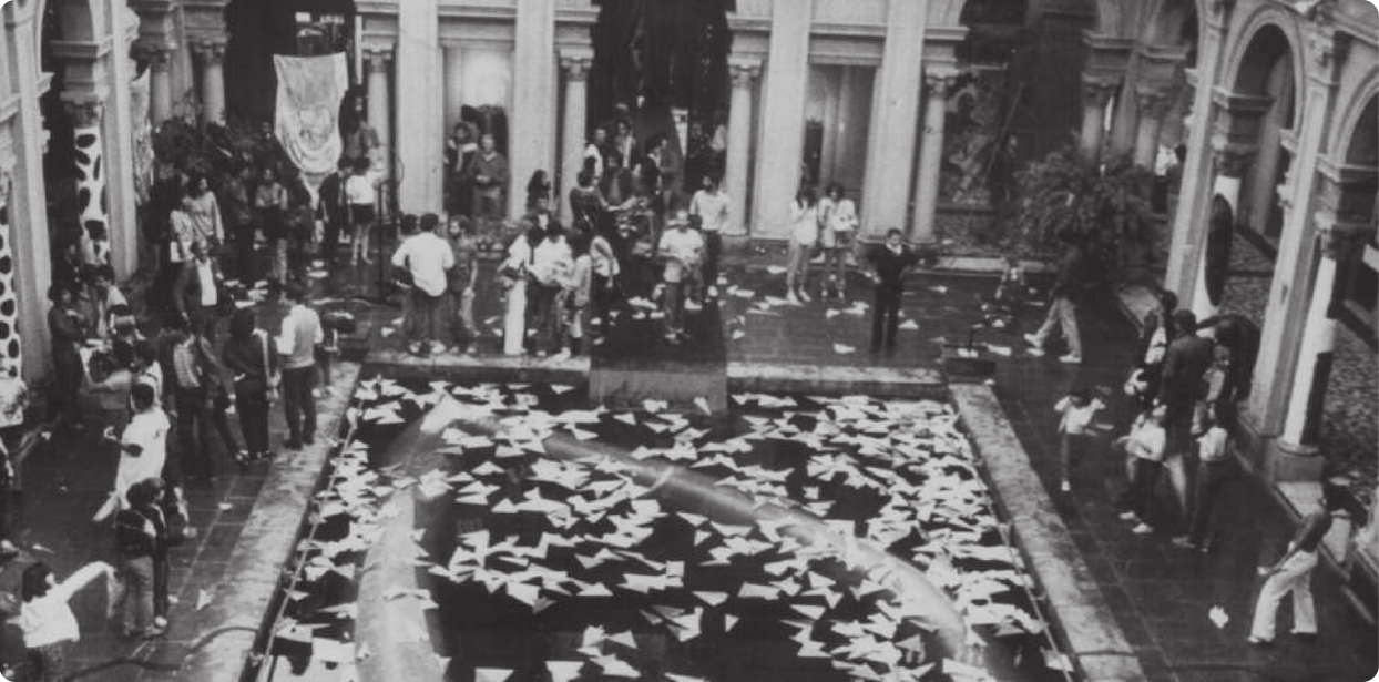 Fotografia em preto e branco de um espelho d'água no centro de um pátio em um edifício de arquitetura neoclássica, com corredores cercados por arcos e colunas. Há muitos de aviões de papel flutuando no espelho d'água, e há grupos de pessoas conversando no pátio.