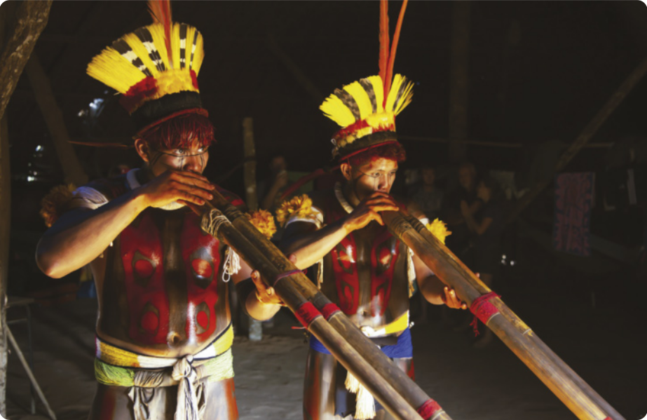 Fotografia de dois homens indígenas com pinturas corporais, cocares e outros adornos tocando flautas longas feitas com dois troncos de bambu. Eles estão sob um telhado de palha, à noite, e ao fundo há pessoas assistindo.