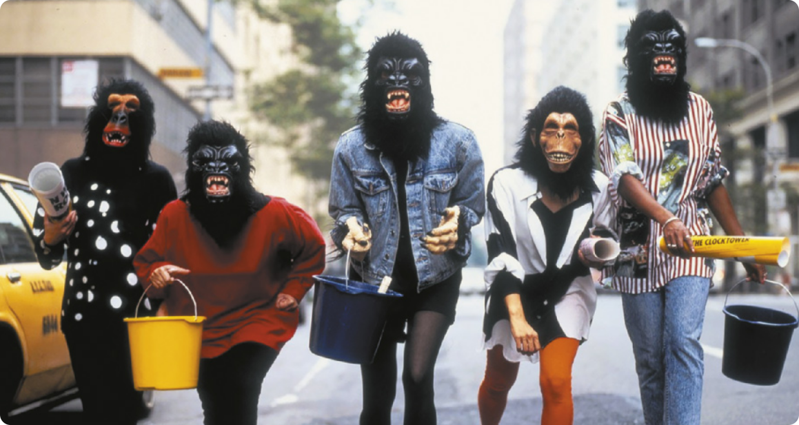 Fotografia de cinco mulheres caminhando em uma via pública, carregando baldes e pôsteres enrolados. Todas elas estão usando máscaras de cabeças de macacos e gorilas.