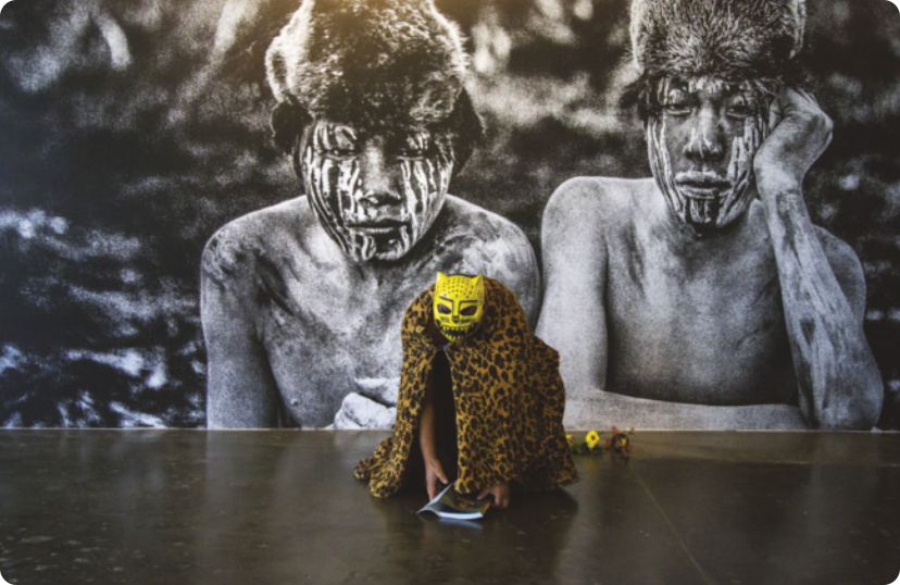 Fotografia de um homem indígena em um espaço expositivo, agachado no chão usando uma fantasia de onça-pintada, composta de uma máscara de onça e um manto de pele de onça. Atrás dele, há uma fotografia em preto e branco com o retrato de dois meninos indígenas com os rostos pintados, usando uma touca de pelo animal.