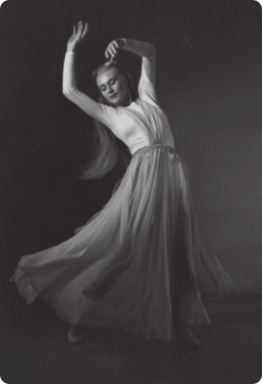 Fotografia de Nina Verchinina. Ela é uma bailarina de cabelos longos, usando uma saia longa. Ela está em pé, com o corpo inclinado para trás e para o lado, com os braços erguidos e os cotovelos levemente flexionados, com as mãos acima da cabeça. Uma das pernas está estendida e a outra está com o joelho flexionado, pisando com a ponta do pé.