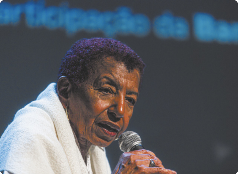 Fotografia de Leci Brandão. Ela é uma mulher idosa com cabelos curtos, usando um casaco, falando com um microfone na mão.