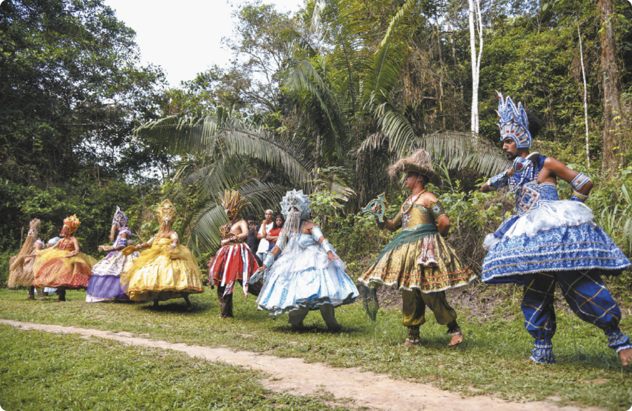 Fotografia de um grupo de pessoas enfileiradas em um gramado, usando saias e trajes festivos representando orixás, com muitos adereços e ornamentos, além de máscaras, chapéus, coroas e capacetes. Cada um deles segura um objeto simbólico referente a um orixá.