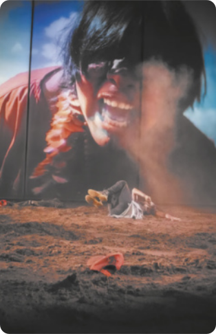 Cena de filme em que uma pessoa está deitada em um chão de terra, com uma grande imagem projetada em uma tela ao fundo, mostrando o rosto de uma mulher indígena gritando, com os cabelos caídos sobre o rosto.