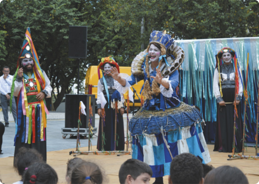 Fotografia de artistas com fantasias e rostos pintados, apresentando uma peça teatral em uma praça, com árvores ao fundo. Há uma atriz com um vestido ricamente adornado, usando uma coroa; um ator com um chapéu pontiagudo e adornado com fitas colorias; e uma mulher com um chapéu colorido ao fundo. Há crianças na plateia e caixas de som em volta dos atores.