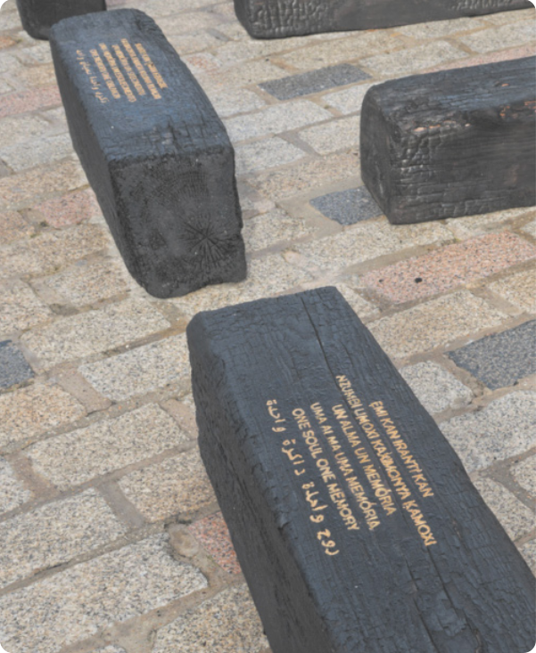 Fotografia de blocos de madeira carbonizada no chão de uma praça. Neles está escrito, em seis línguas, um poema em que um dos versos diz: 'Uma alma uma memória'.