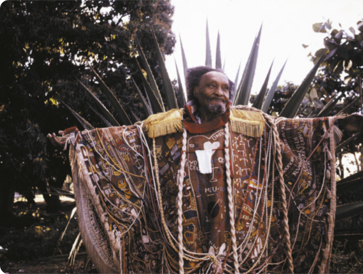 Fotografia de Arthur Bispo do Rosário ao ar livre, com árvores ao fundo. Ele é um homem idoso e calvo, com barba e cabelos grisalhos, de braços abertos. Ele está vestido com um manto ricamente bordado, com diversos tipos de materiais e adornos, além de fios e cordões. Os bordados representam figuras diversas, além de trechos de texto e palavras.