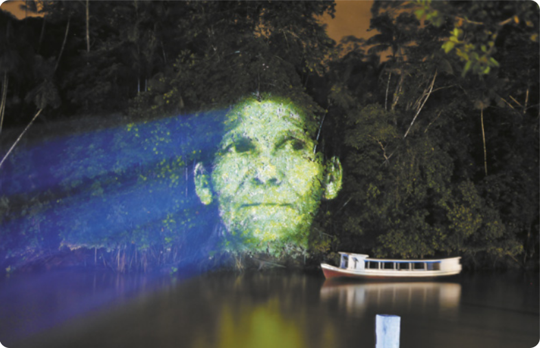Fotografia de árvores à beira de um rio, com um barco flutuando na água. Uma grande imagem luminosa do rosto de uma mulher indígena idosa é projetada sobre as árvores, misturando-se com a textura das folhas e as cores da vegetação.
