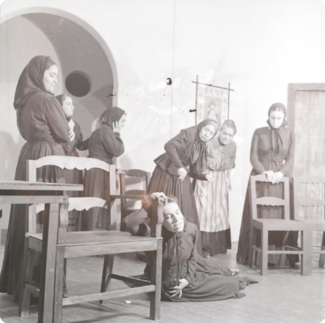 Fotografia em preto e branco de um grupo de mulheres de vestido e lenços na cabeça, em uma sala, atuando em uma cena teatral em que uma delas está deitada no chão, segurando a barriga, com o braço apoiado em uma cadeira, enquanto as outras, em pé, a observam, preocupadas.