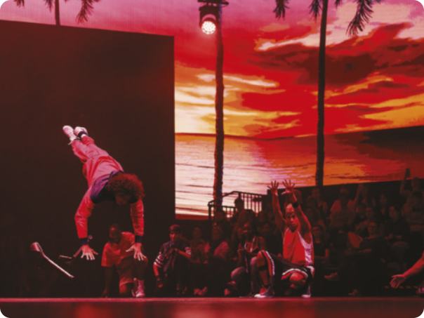 Fotografia de Youcef Mecheri. Ele é um dançarino de breaking em um palco, realizando um movimento acrobático com o corpo no ar, de cabeça para baixo, com as mãos em direção ao chão e as pernas estendidas em diagonal para o alto. Ao lado dele está um homem agachado que estende os braços para o alto na direção de Youcef Mecheri.