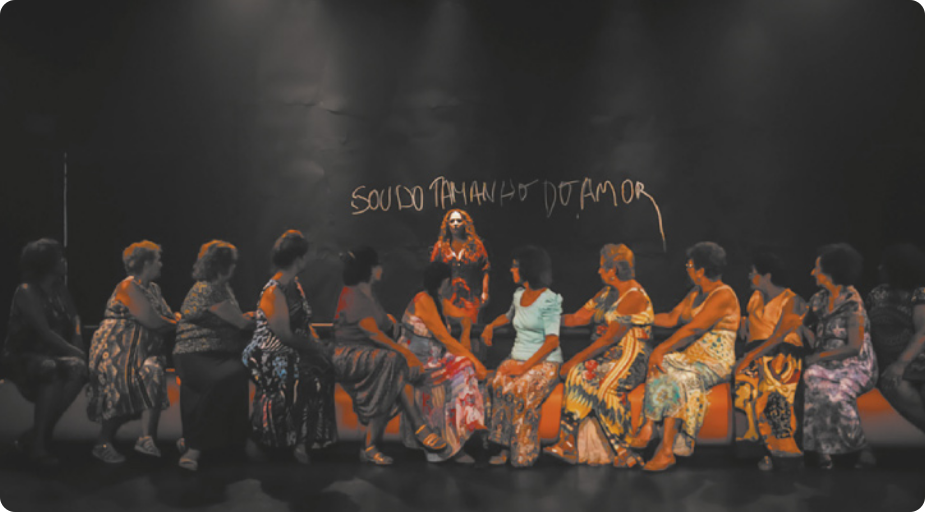 Fotografia de um grupo de mulheres sentadas lado a lado em um banco sobre um palco. Elas usam vestidos e olham para trás, onde está uma mulher em pé. Ao fundo, está escrita a frase 'sou do tamanho do amor' em uma parede.