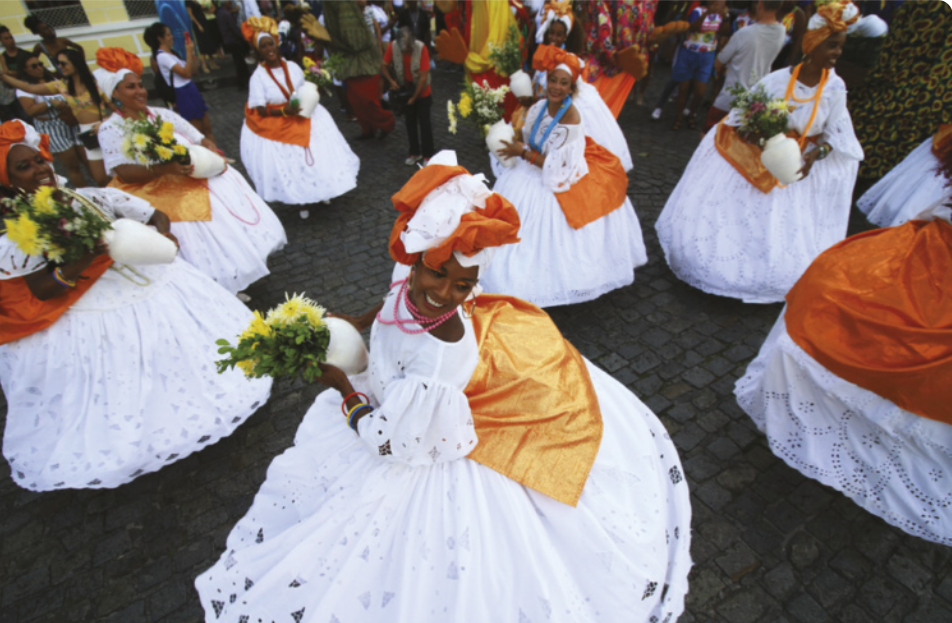 Fotografia de um grupo de mulheres uniformizadas com vestidos brancos, colares e lenços na cabeça, segurando jarros com buquês de flores. Elas estão dançando em uma rua, em movimentos circulares, com as saias esvoaçantes.