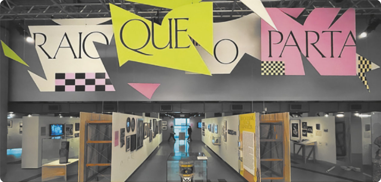 Fotografia de uma exposição com um painel na fachada com as palavras 'Raio que o parta' dispostas em painéis com formas abstratas e geométricas distribuídas em planos e profundidades diversas.