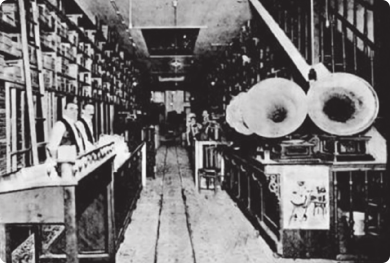 Fotografia em preto e branco de uma loja com vitrolas expostas sobre uma bancada à direita, com dois homens atrás de um balcão à esquerda, em frente a uma estante alta repleta de discos.