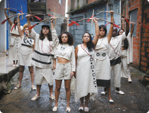 Fotografia de um grupo de homens e mulheres enfileirados em pé em uma viela, usando roupas brancas, com pinturas de inspiração indígena no rosto, erguendo arco e flechas para o alto.
