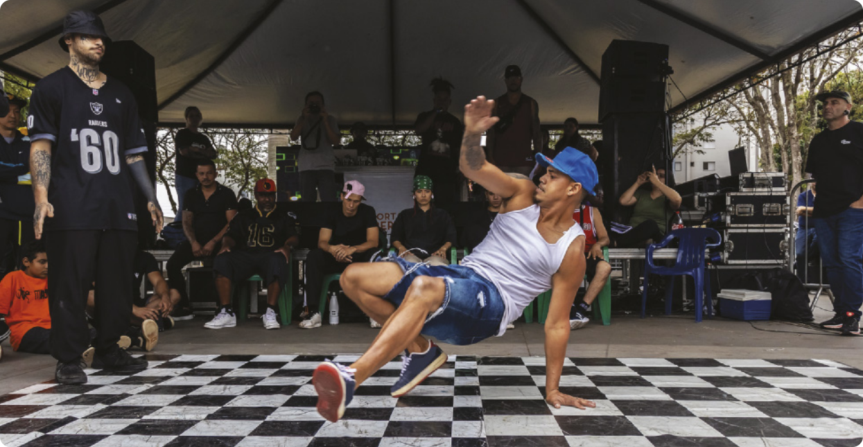 Fotografia de um homem de boné realizando um movimento de dança agachado no chão, apoiado com a mão e a ponta de um pé no chão, estendendo a outra perna à frente, com um braço erguido acima da cabeça. Em volta dele há um grupo de pessoas sentadas e em pé observando a apresentação do homem.