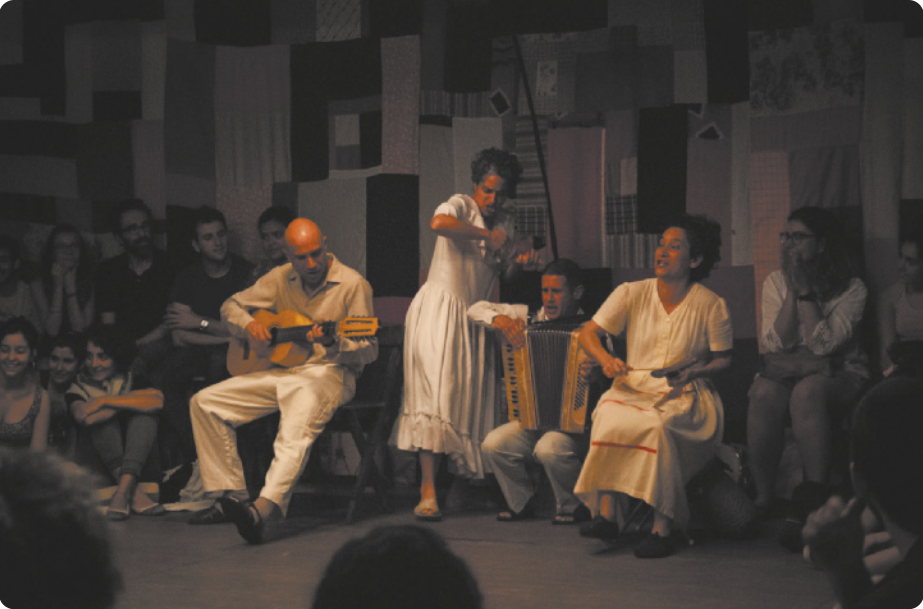 Fotografia de artistas se apresentando em uma sala, com o público sentado em torno deles. Há um homem sentado em uma cadeira tocando uma viola caipira; um homem sentado tocando acordeon; uma mulher em pé tocando um instrumento de percussão; e uma mulher sentada cantando e tocando um reco-reco.
