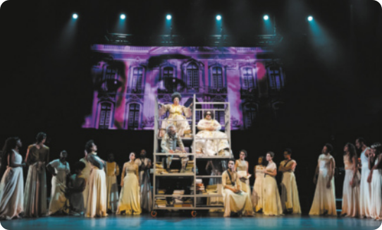 Fotografia de um grupo de atores e atrizes em um palco, vestidos com roupas brancas. No centro do palco, há um andaime onde estão sentadas duas mulheres de vestido e um homem. Ao fundo há uma imagem cenográfica da fachada de um edifício, com a projeção de olhos de uma pessoa sobre a fachada.
