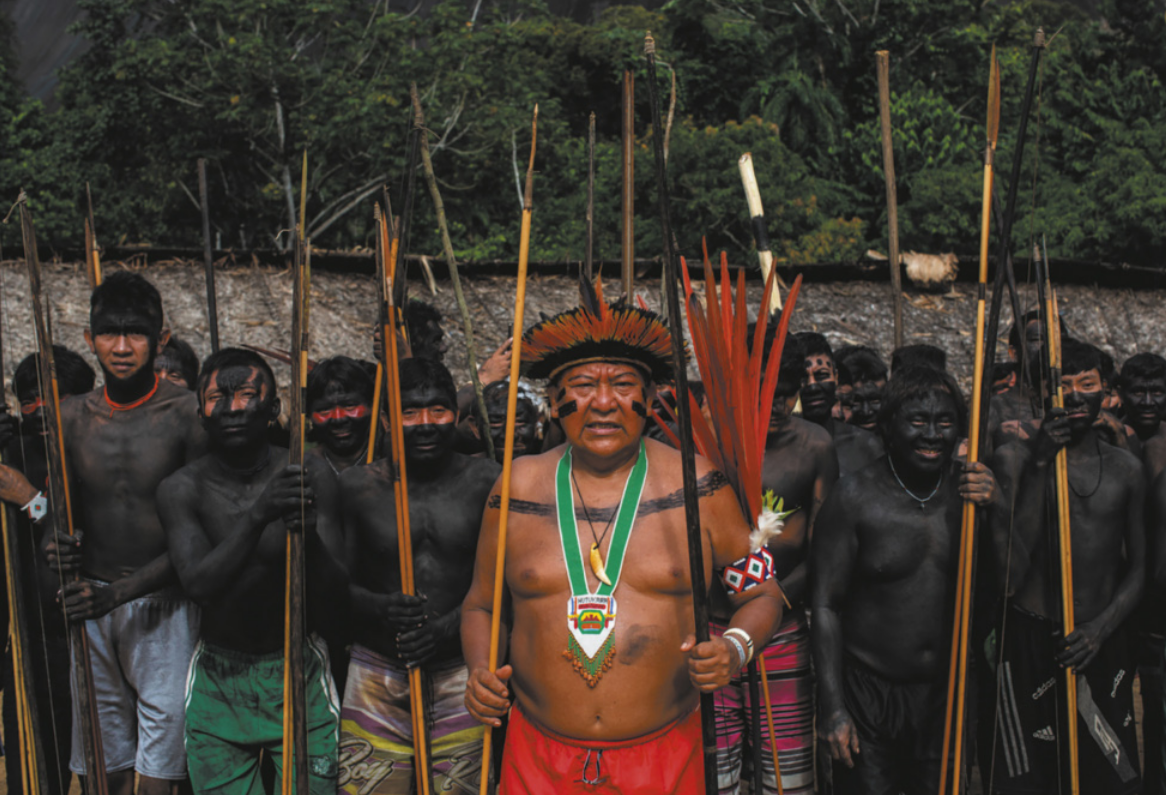Fotografia de Davi Kopenawa: um homem indígena com pinturas corporais, colares, adornos com penas e um cocar, segurando um arco e flecha. Ele está à frente de um grupo de homens indígenas, todos com os corpos pintados de preto, segurando arcos e flechas.