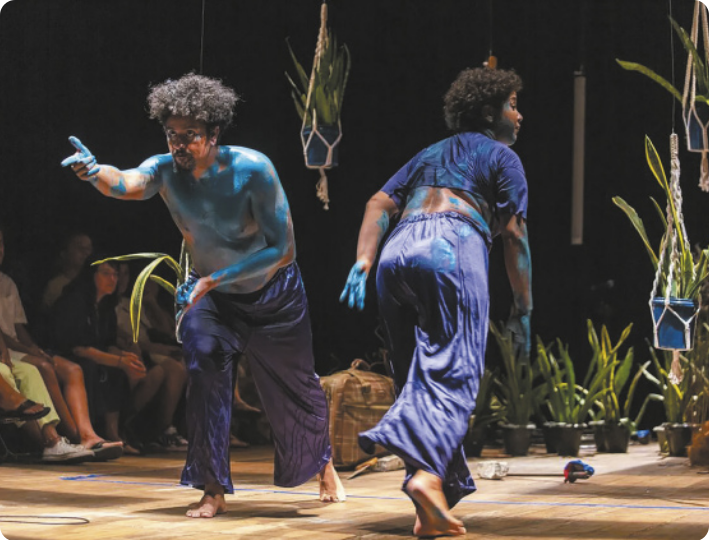 Fotografia de um homem e uma mulher em um palco com diversas plantas em vasos no cenário. Eles estão com os corpos pintados de tinta azul e inclinados para a frente, com a perna da frente flexionada e a perna de trás estendida, pisando com a ponta do pé. Eles estão lado a lado e se inclinam em sentidos opostos, e o homem está apontando o dedo indicador para a frente.