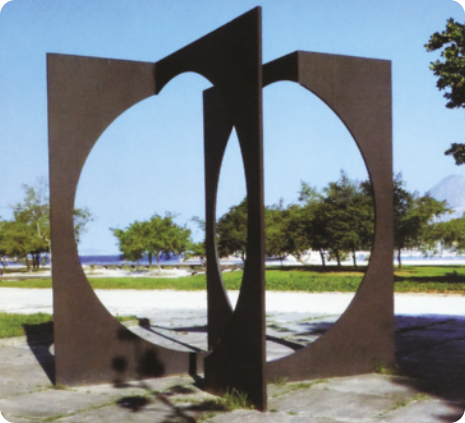 Fotografia de escultura metálica de formas geométricas abstratas em um espaço aberto, com uma praia ao fundo. A escultura consiste em duas formas quadradas com círculos vazados inscritos dentro dos quadrados, dispostas de modo que criam dois planos ortogonais entre si, que se interpõem e se articulam à medida que o observador se movimenta em torno da escultura, configurando formas semicirculares e elípticas diversas nos espaços vazados.