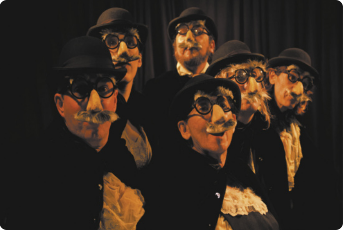 Fotografia de um grupo de seis atores usando chapéu coco e óculos de armação circular, com narizes e bigodes grisalhos postiços.