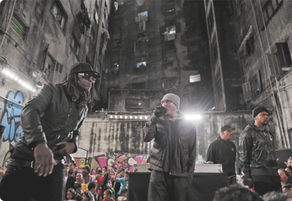 Fotografia de dois homens em um palco à noite, em meio a edifícios com a pintura degradada. Um deles está de óculos escuros e boné, com tranças no cabelo e uma jaqueta de couro, segurando um microfone; e o outro está de touca na cabeça e luvas, usando um casaco, cantando com um microfone na mão.