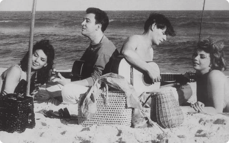 Fotografia em preto e branco de dois homens tocando violão, um de costas para o outro, e duas mulheres deitadas ao lado deles, em uma praia. Os homens tocando violão são João Gilberto, à esquerda, vestido com uma camisa; e Tom Jobim, à direita, sem camiseta.