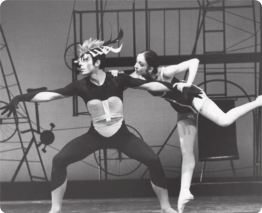 Fotografia em preto e branco de John Neumeier: um bailarino com um adereço com penas sobre a cabeça; e Marcia Haydée: uma bailarina dançando apoiada na ponta de um pé, inclinando o corpo e apoiando as mãos em John Neumeier.