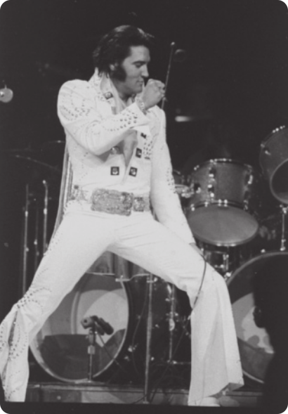 Fotografia em preto e branco de Elvis Presley. Ele é um homem de cabelos curtos, com topete e costeletas, usando jaqueta e calças brancas enfeitadas com adornos brilhantes, com as pernas afastadas e os joelhos flexionados, cantando com um microfone na mão em um palco.