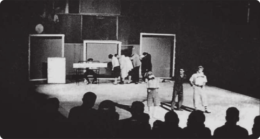 Fotografia em preto e branco de atores em um palco. Há três homens em pé diante da plateia, e ao fundo está um homem sentado atrás de uma espécie de mesa, ao lado de um grupo de homens e mulheres em fila, com as cabeças curvadas em direção ao ator que está à frente na fila.