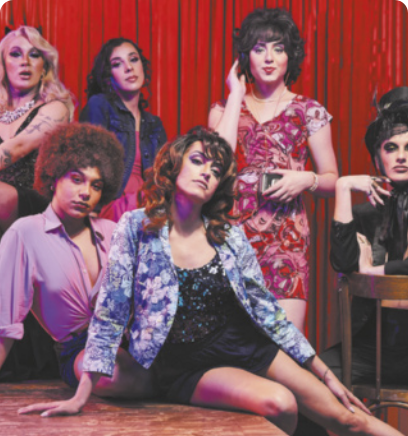 Fotografia de um grupo de seis travestis usando vestidos, jaquetas, maquiagens e penteados diversos, uma delas com um chapéu, posando para a fotografia em frente a uma cortina vermelha.