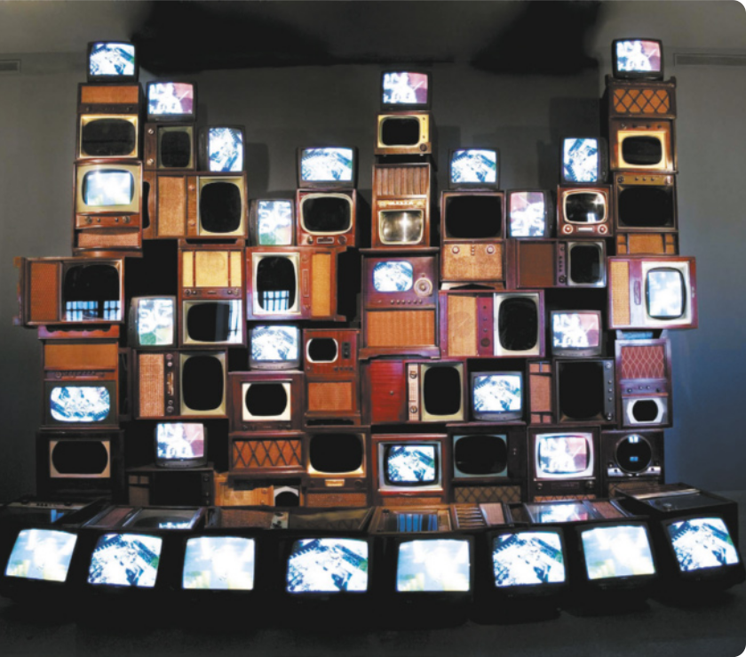 Fotografia de instalação com muitos televisores empilhados, com aparências e formatos diversos. Alguns deles estão ligados, mostrando imagens na tela.