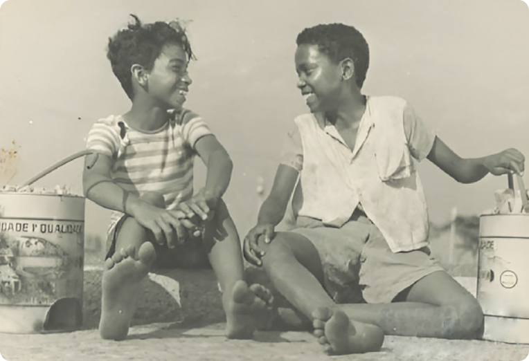 Fotografia em preto e branco de dois meninos descalços, sentados em uma calçada, conversando, cada um deles portando uma grande lata com embalagens de amendoim para vender.