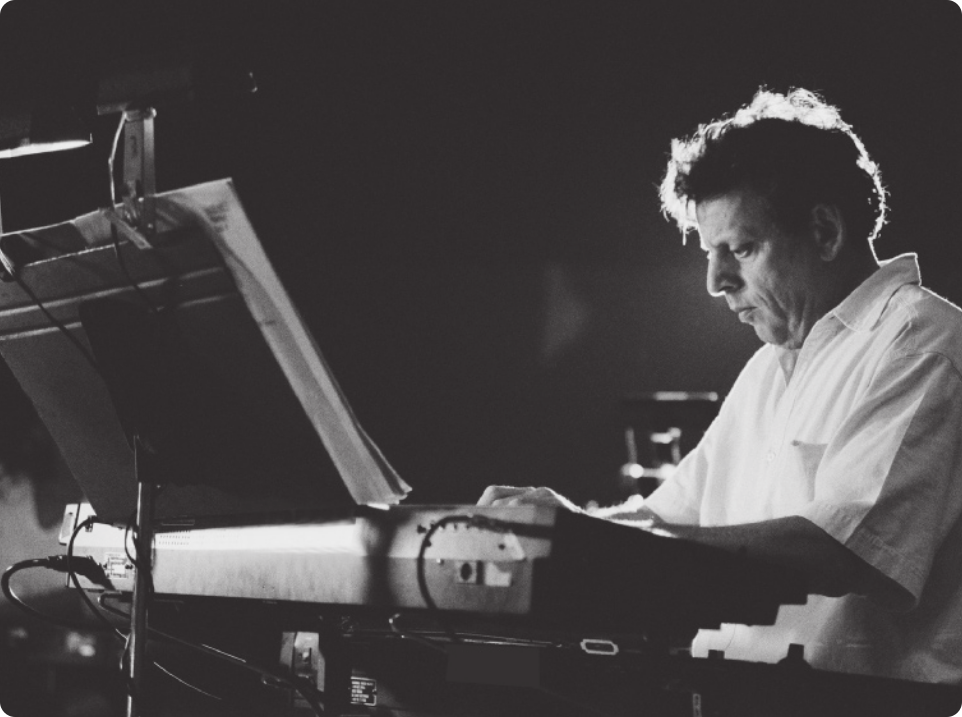 Fotografia em preto e branco de Philip Glass. Ele é um homem de cabelos curtos, usando uma camisa, tocando um teclado eletrônico.